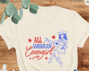 May include: A beige t-shirt with a graphic of a woman playing a guitar. The text "ALL AMERICAN Cowgirl" is printed on the shirt in red and blue.