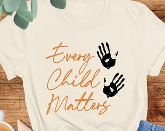 May include: Cream-coloured t-shirt with the text "Every Child Matters" in orange script, accompanied by two black handprints. The shirt is paired with blue jeans and blush-coloured shoes. A cup of coffee with latte art is also visible.