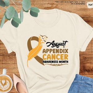 May include: A beige t-shirt with a brown ribbon and a yellow feather. The text on the shirt reads "August is Appendix Cancer Awareness Month".
