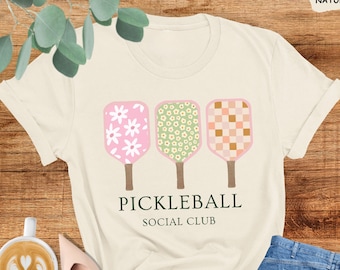 Pode incluir: Uma camiseta branca com um design de raquete de pickleball verde, rosa e laranja. O texto "PICKLEBALL SOCIAL CLUB" está impresso abaixo das raquetes.