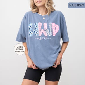 May include: A blue jean colored t-shirt with the word "WILD" in a floral and gingham pattern. The shirt has short sleeves and a relaxed fit. A small bow graphic is below the word.