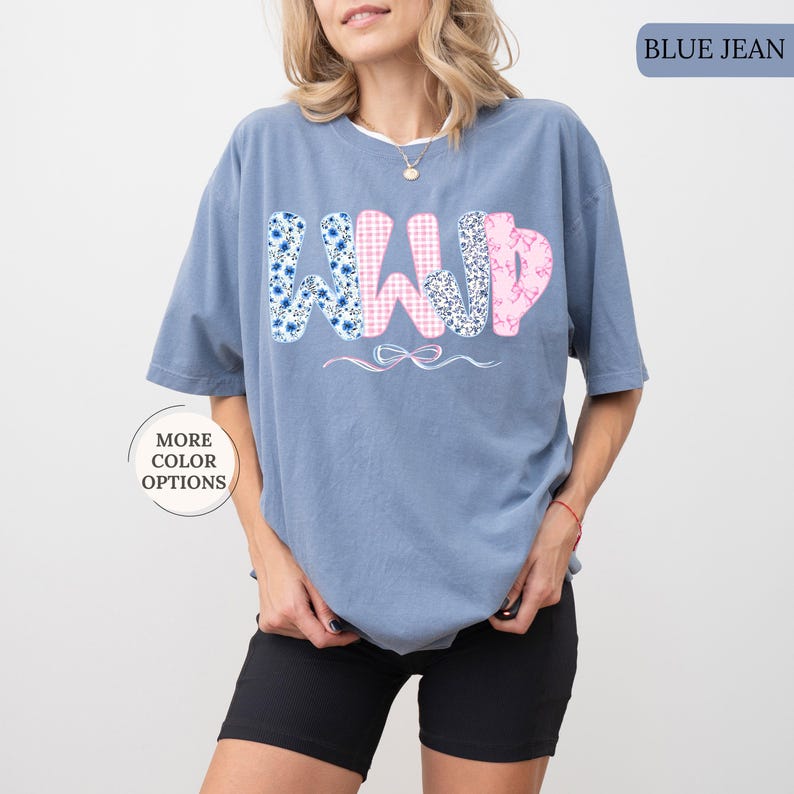 May include: A blue jean colored t-shirt with the word "WILD" in a floral and gingham pattern. The shirt has short sleeves and a relaxed fit. A small bow graphic is below the word.