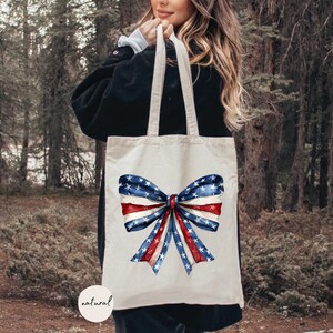 May include: A white canvas tote bag with a red, white, and blue bow printed on it. The bow is made of ribbon with stars and stripes. The bag has a natural fabric label.