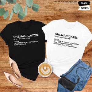 Pode incluir: Duas camisetas pretas e brancas com uma defini&ccedil;&atilde;o das palavras "Shenanigator" e "Shenanigatee". As camisetas est&atilde;o dispostas sobre uma superf&iacute;cie de madeira com uma x&iacute;cara de caf&eacute;, um par de sapatos e uma bolsa.