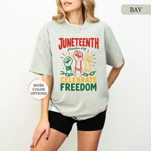 K&ouml;nnte beinhalten: Hellgraues T-Shirt mit den Worten "Juneteenth freedom day" in Rot und "Celebrate Freedom" in Gr&uuml;n. Das Design zeigt drei erhobene F&auml;uste in Gr&uuml;n, Rot und Gelb mit Ketten darunter.