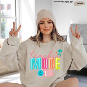 May include: A light beige sweatshirt with the text "teacher mode off" in colorful letters. The text is surrounded by a colorful light bulb and palm trees.