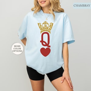 May include: A light blue t-shirt with a gold glitter crown and a red heart with the letter "Q" in the center. The text "CHAMBRAY" is in the upper right corner of the image.