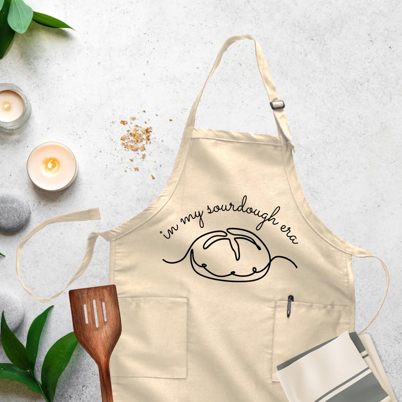 May include: A beige kitchen apron with a black outline of a sourdough loaf and the text 'in my sourdough era'.