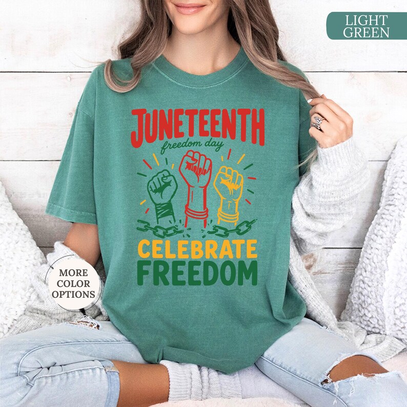 K&ouml;nnte beinhalten: Hellgr&uuml;nes T-Shirt mit den roten Worten "Juneteenth freedom day" und dem gr&uuml;nen Satz "Celebrate Freedom". Das Shirt zeigt drei erhobene F&auml;uste in Gr&uuml;n, Rot und Gelb, die durch Ketten verbunden sind.