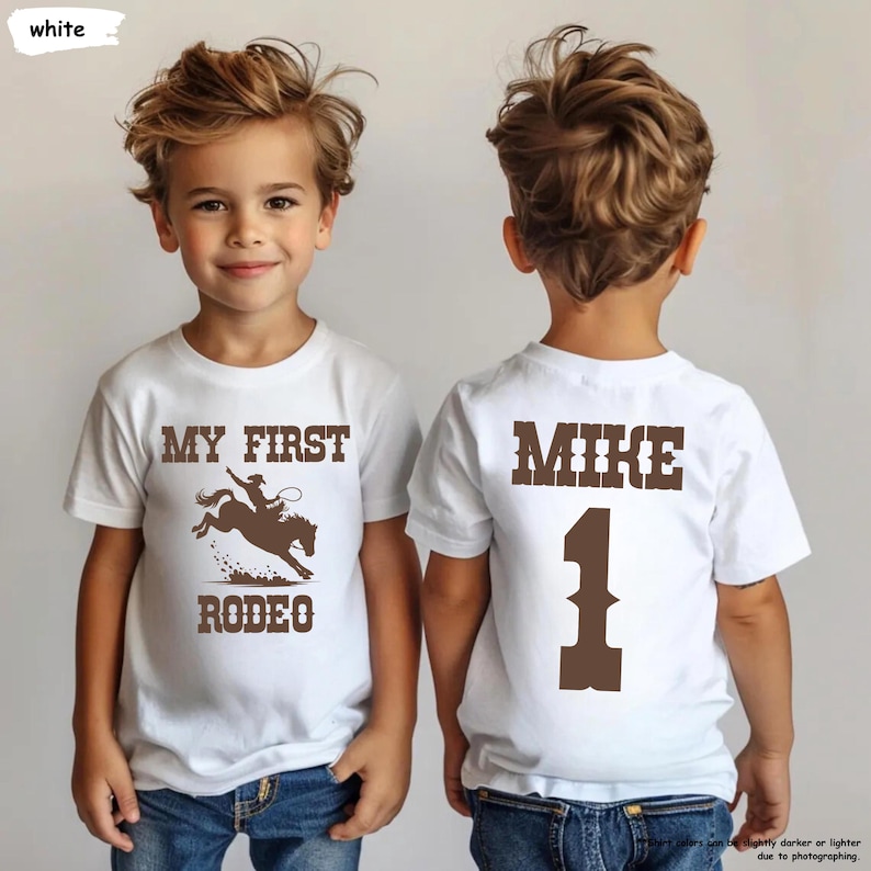 May include: White t-shirt with a brown graphic of a rodeo rider on a horse, the text "MY FIRST RODEO" is above and below the graphic. The back of the shirt has the name "MIKE" and the number "1" in brown.