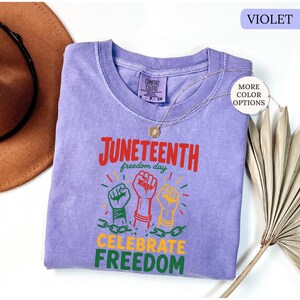 K&ouml;nnte beinhalten: Ein violettes T-Shirt mit dem Schriftzug "Juneteenth freedom day" in Rot und Gr&uuml;n. Darunter befinden sich drei erhobene F&auml;uste in Gr&uuml;n, Rot und Gelb, mit den Worten "Celebrate Freedom" in Gelb und Gr&uuml;n. Eine goldene Halskette liegt auf dem T-Shirt.