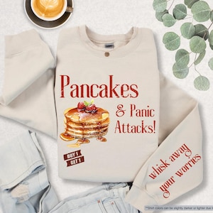 May include: A beige sweatshirt with the text "Pancakes & Panic Attacks!" in red. A stack of pancakes with strawberries is illustrated next to the text. The text "Buy 1 Get 1" is in a red rectangle below the pancakes. The text "Whisk away your worries" is in red on the right sleeve.