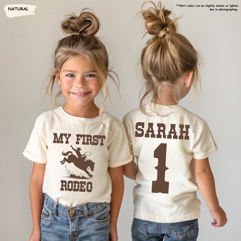May include: Two young girls wearing matching cream-colored t-shirts and blue jeans. The front of the shirt says "MY FIRST RODEO" with a rodeo graphic. The back of the shirt says "SARAH" and the number "1".