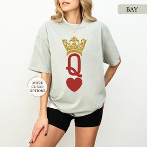 May include: A light gray t-shirt with a red heart and a gold glitter crown above the letter "Q". The text "BAY" is in the upper right corner of the image.