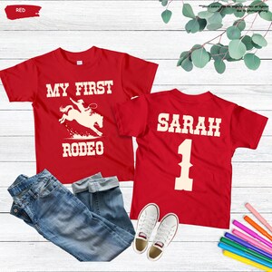 May include: Two red t-shirts with white text and graphics. One shirt says "MY FIRST RODEO" with a rodeo rider graphic. The other shirt says "SARAH 1". Blue jeans and white sneakers are also in the image.
