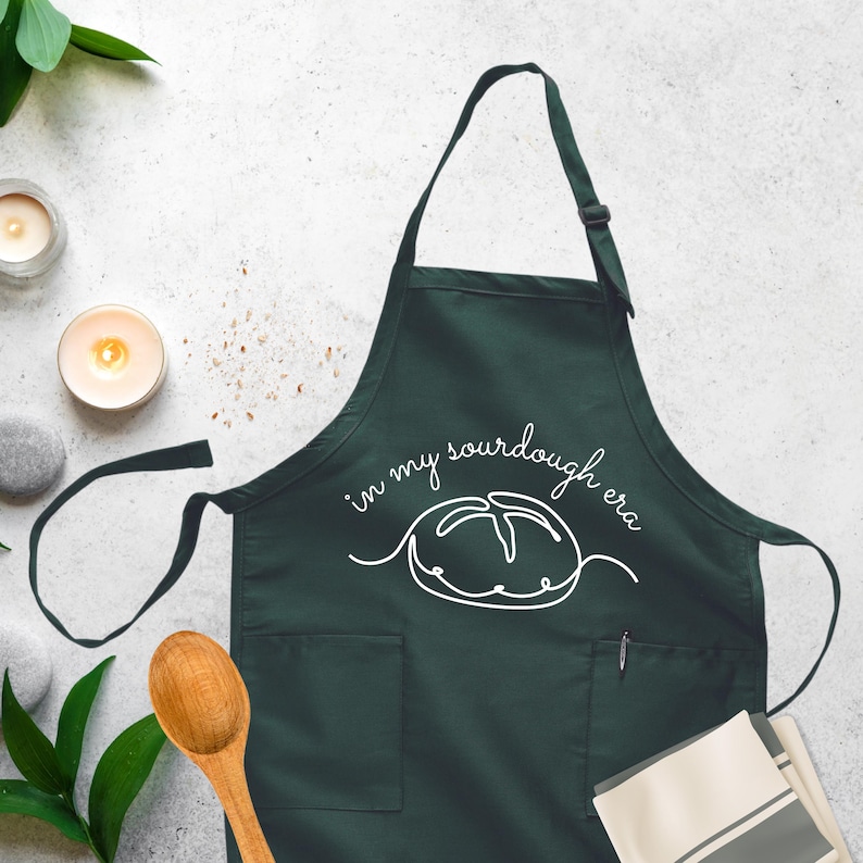 Puede incluir: Un delantal verde oscuro con un contorno blanco de una hogaza de pan de masa madre y el texto "in my sourdough era".
