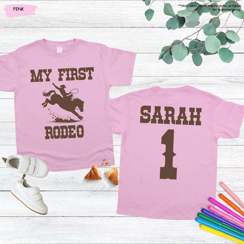 May include: Pink t-shirts with brown text and graphics. One shirt says "MY FIRST RODEO" with a rodeo rider graphic. The other shirt says "SARAH" and the number "1". White sneakers and colorful markers are also visible.