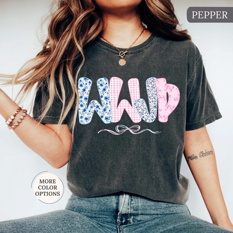 May include: Dark gray t-shirt with the letters "WWJD" in a floral, gingham, and marble pattern. The shirt has a relaxed fit and a small bow graphic below the letters. The word "Pepper" is in the top right corner.