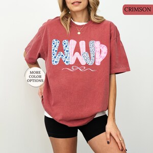 May include: Crimson-colored t-shirt with the letters "WWJD" in a floral, gingham, and patterned design. The shirt has short sleeves and a relaxed fit. The text "MORE COLOR OPTIONS" is on the left side of the shirt.