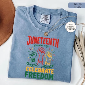K&ouml;nnte beinhalten: Ein blaues Jeans-T-Shirt mit den Worten "Juneteenth freedom day" und "Celebrate Freedom" in Rot, Gr&uuml;n und Gelb. Das Shirt zeigt drei erhobene F&auml;uste in Gr&uuml;n, Rot und Gelb mit Ketten. Eine goldene Halskette ist &uuml;ber dem Shirt drapiert.