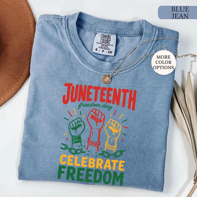 K&ouml;nnte beinhalten: Ein blaues Jeans-T-Shirt mit den Worten "Juneteenth freedom day" und "Celebrate Freedom" in Rot, Gr&uuml;n und Gelb. Das Shirt zeigt drei erhobene F&auml;uste in Gr&uuml;n, Rot und Gelb mit Ketten. Eine goldene Halskette ist &uuml;ber dem Shirt drapiert.