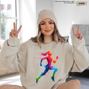 May include: A beige sweatshirt with a colorful silhouette of a woman running. The design features splashes of paint in shades of red, orange, yellow, green, blue, and purple.