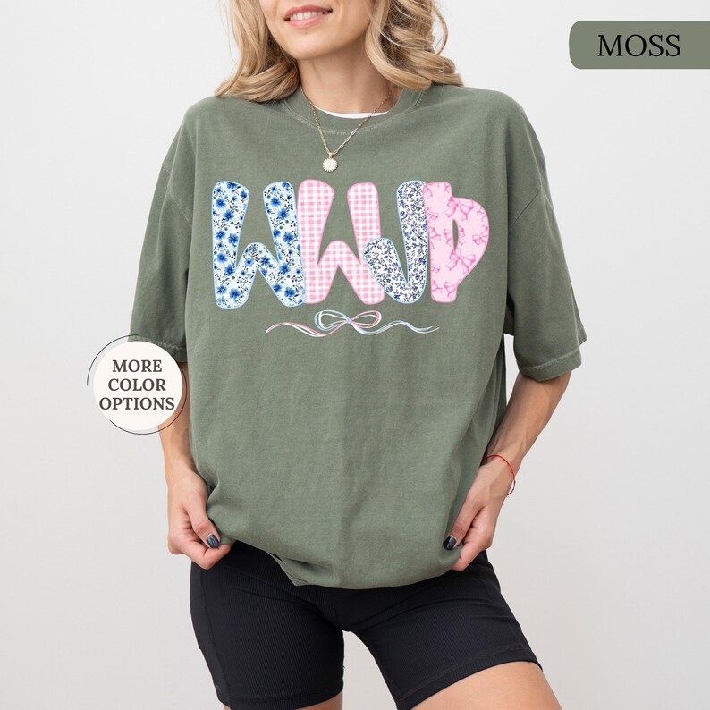 May include: A moss green t-shirt with the letters "WWYD" in a floral and gingham pattern. The "W"s are blue floral, the "W" is pink gingham, and the "D" is pink floral. The shirt has a relaxed fit.