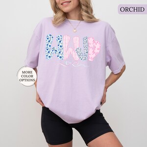 May include: Orchid-colored t-shirt with the word "WILD" in a floral and gingham pattern. The "W" is blue with white flowers, the "I" is pink gingham, the "L" is pink with a floral pattern, and the "D" is blue with white flowers. The shirt is paired with black shorts.