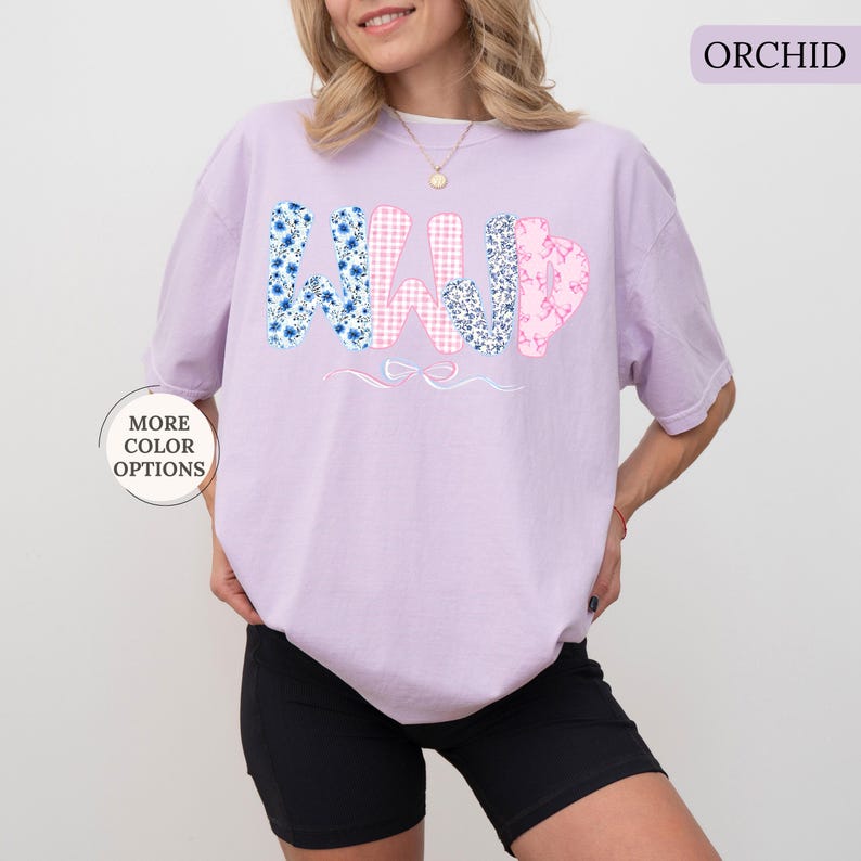 May include: Orchid-colored t-shirt with the word "WILD" in a floral and gingham pattern. The "W" is blue with white flowers, the "I" is pink gingham, the "L" is pink with a floral pattern, and the "D" is blue with white flowers. The shirt is paired with black shorts.