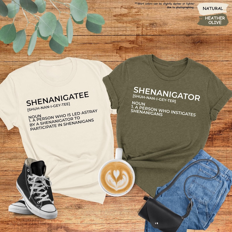 Pode incluir: Duas camisetas com a palavra "Shenanigatee" e "Shenanigator" definida como um substantivo. As camisetas est&atilde;o sobre uma superf&iacute;cie de madeira com uma x&iacute;cara de caf&eacute;, um par de t&ecirc;nis pretos, uma cal&ccedil;a jeans azul e uma bolsa preta.