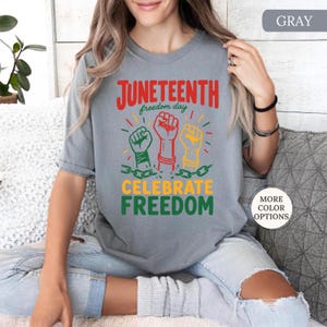 K&ouml;nnte beinhalten: Graues T-Shirt mit den Worten "Juneteenth freedom day" in Rot und Gr&uuml;n. Das Shirt zeigt drei erhobene F&auml;uste in Gr&uuml;n, Rot und Gelb, mit den Worten "Celebrate Freedom" darunter.