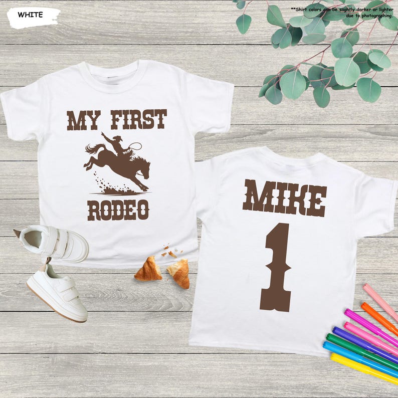 May include: White t-shirts with brown text and graphics. One shirt says "MY FIRST RODEO" with a cowboy on a bucking bronco. The other shirt says "MIKE" and the number "1".