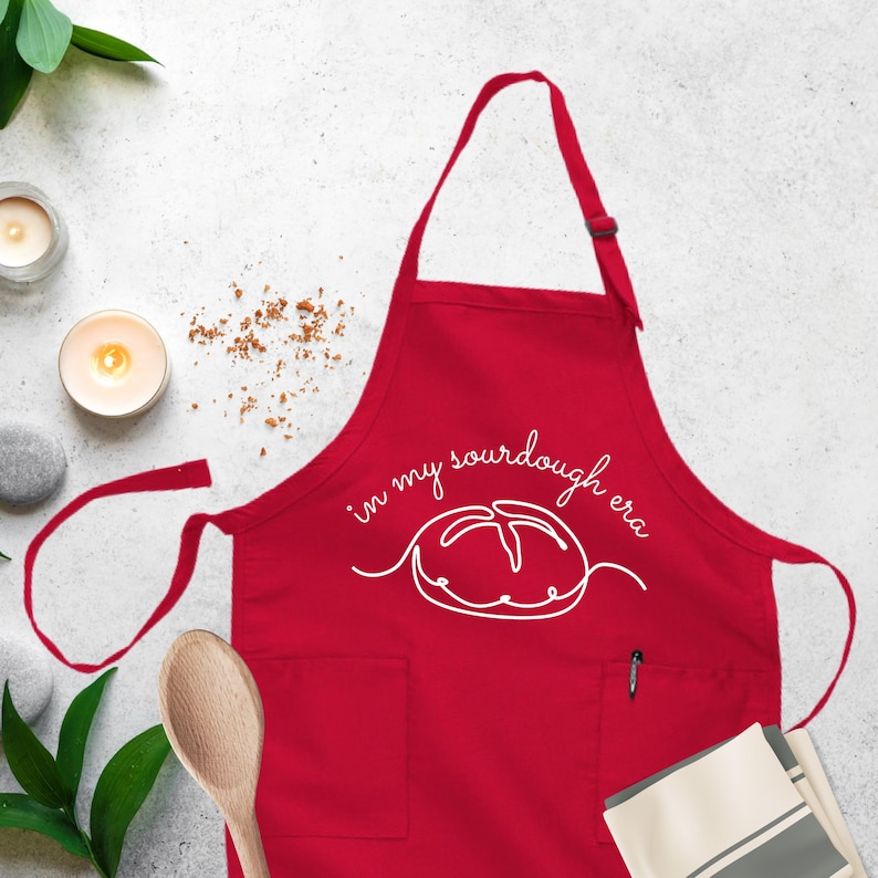 Puede incluir: Delantal de cocina rojo con un contorno blanco de una hogaza de pan de masa madre y el texto "in my sourdough era".