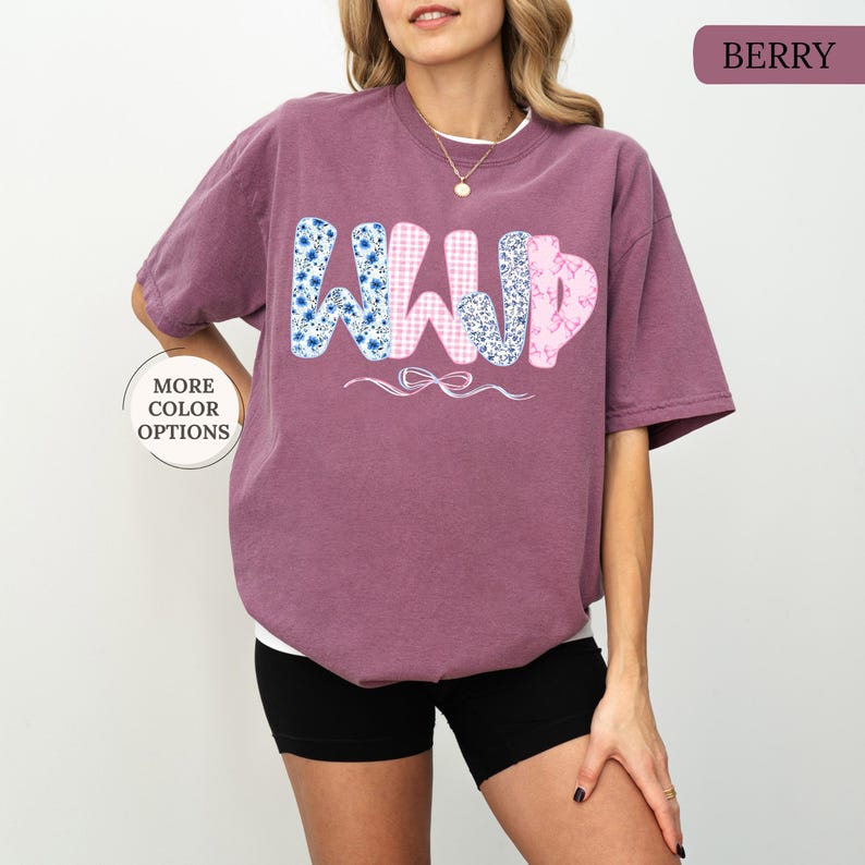 May include: Berry-colored t-shirt with the letters "WWYD" in a playful font. The "W"s are decorated with blue floral patterns, the "W" is pink gingham, and the "D" is a pink floral pattern. A white bow is below the letters.