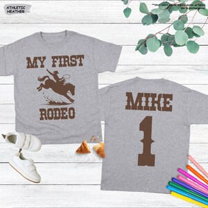 May include: Two gray t-shirts. The front of one shirt says "MY FIRST RODEO" with a graphic of a cowboy on a bucking bronco. The back of the other shirt says "MIKE" and the number "1".