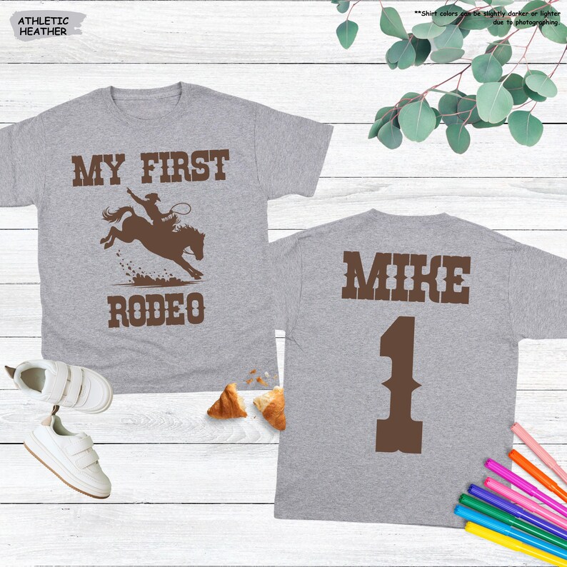 May include: Two gray t-shirts. The front of one shirt says "MY FIRST RODEO" with a graphic of a cowboy on a bucking bronco. The back of the other shirt says "MIKE" and the number "1".