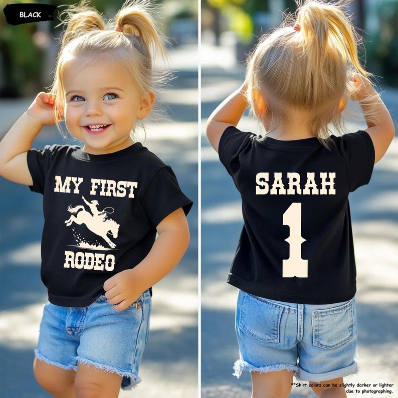 May include: Black toddler t-shirt with "MY FIRST RODEO" and a rodeo graphic on the front. The back of the shirt has the name "SARAH" and the number "1". The child is wearing denim shorts.