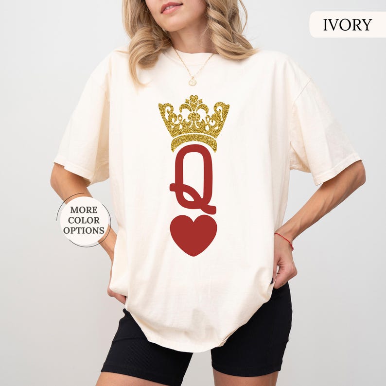 May include: A white t-shirt with a gold glitter crown and a red heart with a gold glitter "Q" in the center. The shirt is available in ivory.