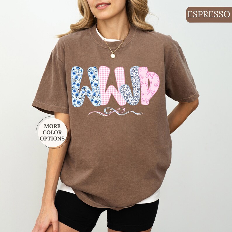 May include: Espresso-colored t-shirt with the letters "WWD" in a floral and gingham pattern. The shirt has a relaxed fit and short sleeves. A small bow design is below the letters. The words "More Color Options" are in a circle on the left.