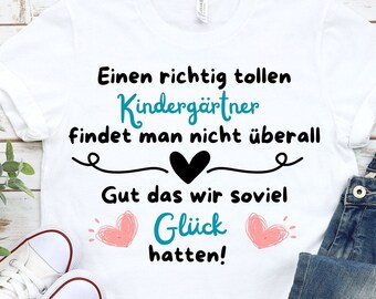 May include: White t-shirt with a blue and black graphic that says "Einen richtig tollen Kindergärtner findet man nicht überall Gut das wir soviel Glück hatten!" with two pink hearts on either side of the word "Glück".