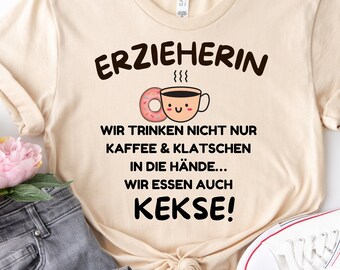 May include: A beige t-shirt with the text "ERZIEHERIN" and a cartoon image of a coffee cup with a donut. The text below the image reads "WIR TRINKEN NICHT NUR KAFFEE & KLATSCHEN IN DIE HÄNDE... WIR ESSEN AUCH KEKSE!"