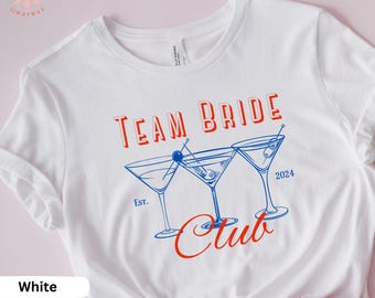 Może przedstawiać: Biała koszulka z napisem "Team Bride Club" w kolorze czerwonym i niebieskim. Tekst jest otoczony trzema szklankami martini z oliwkami. Na koszulce nadrukowano również rok "2024" i "Est."
