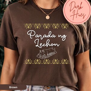 May include: Brown t-shirt with the text "Parada ng Lechon" and a graphic of a roasted pig. The design includes decorative floral elements in gold. The shirt is a crew neck style.