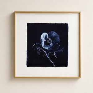 May include: A square framed wall art piece featuring a dark navy blue cyanotype print of a single flower. The artwork displays a white, negative-style floral image with a central bloom and stem set against a deep, solid indigo background.