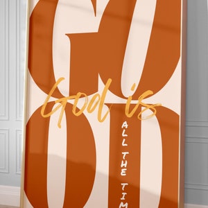 Könnte beinhalten: Ein gerahmter Druck mit den Worten "Gott ist gut" in goldener Schrift und "die ganze Zeit" in kleinerer goldener Schrift. Der Hintergrund ist pfirsichfarben mit großen orangefarbenen Buchstaben, die das Wort "GUT" bilden.