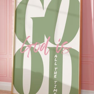 May include: A framed art print with the words "God is Good All the Time" in a large, stylized font. The word "GOOD" is in large, olive green ovals, with the words "God is" in pink script. The words "ALL THE TIME" are in vertical, olive green text.