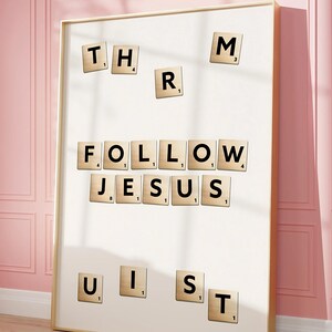 Puede incluir: Una obra de arte enmarcada con las palabras "FOLLOW JESUS" escritas con fichas de Scrabble. Fichas adicionales deletrean "THR", "M", "R", "UIST". La obra de arte está sobre un fondo blanco y enmarcada en un marco de madera clara.