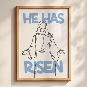 Puede incluir: Una impresión artística enmarcada con un dibujo lineal minimalista de una figura con los brazos extendidos. Las palabras "HE HAS RISEN" están impresas en azul claro encima y debajo de la figura. La obra de arte está sobre un fondo crema.