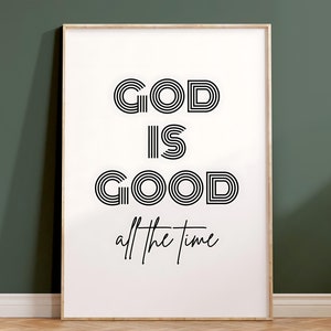 Puede incluir: Una impresión enmarcada con el texto "GOD IS GOOD all the time" en un estilo de fuente retro en blanco y negro. La impresión está sobre un fondo blanco y está enmarcada en un marco de madera claro.
