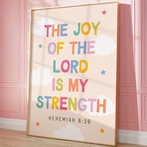 Puede incluir: Un cuadro enmarcado con las palabras "THE JOY OF THE LORD IS MY STRENGTH" en letras coloridas y divertidas. La impresión incluye ilustraciones de nubes y estrellas, y el versículo bíblico Nehemías 8:10. El marco es dorado.